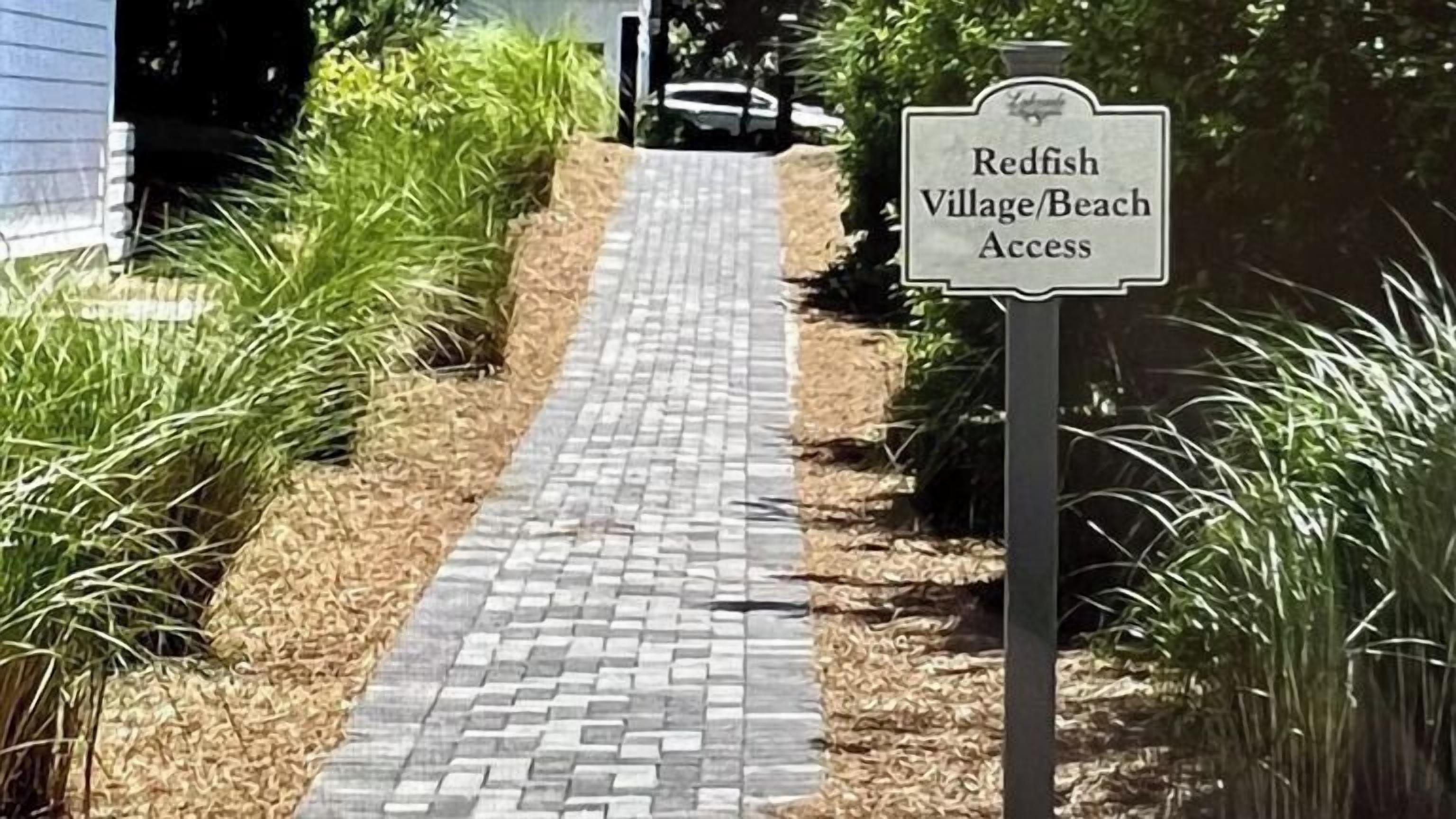 A paved pathway leading to Redfish Village Beach Access, surrounded by grass and shrubs.