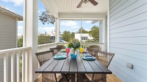 Een overdekte eethoek buiten met een houten tafel en rieten stoelen, een plafondventilator en uitzicht op omliggende huizen.