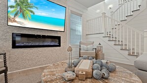 A living room with a large TV displaying a beach scene, a fireplace, a staircase, and a round coffee table with decorative items. - Steps to Beach Pool, Bikes & Game Room Seacrest 5BR (Panama City Beach)