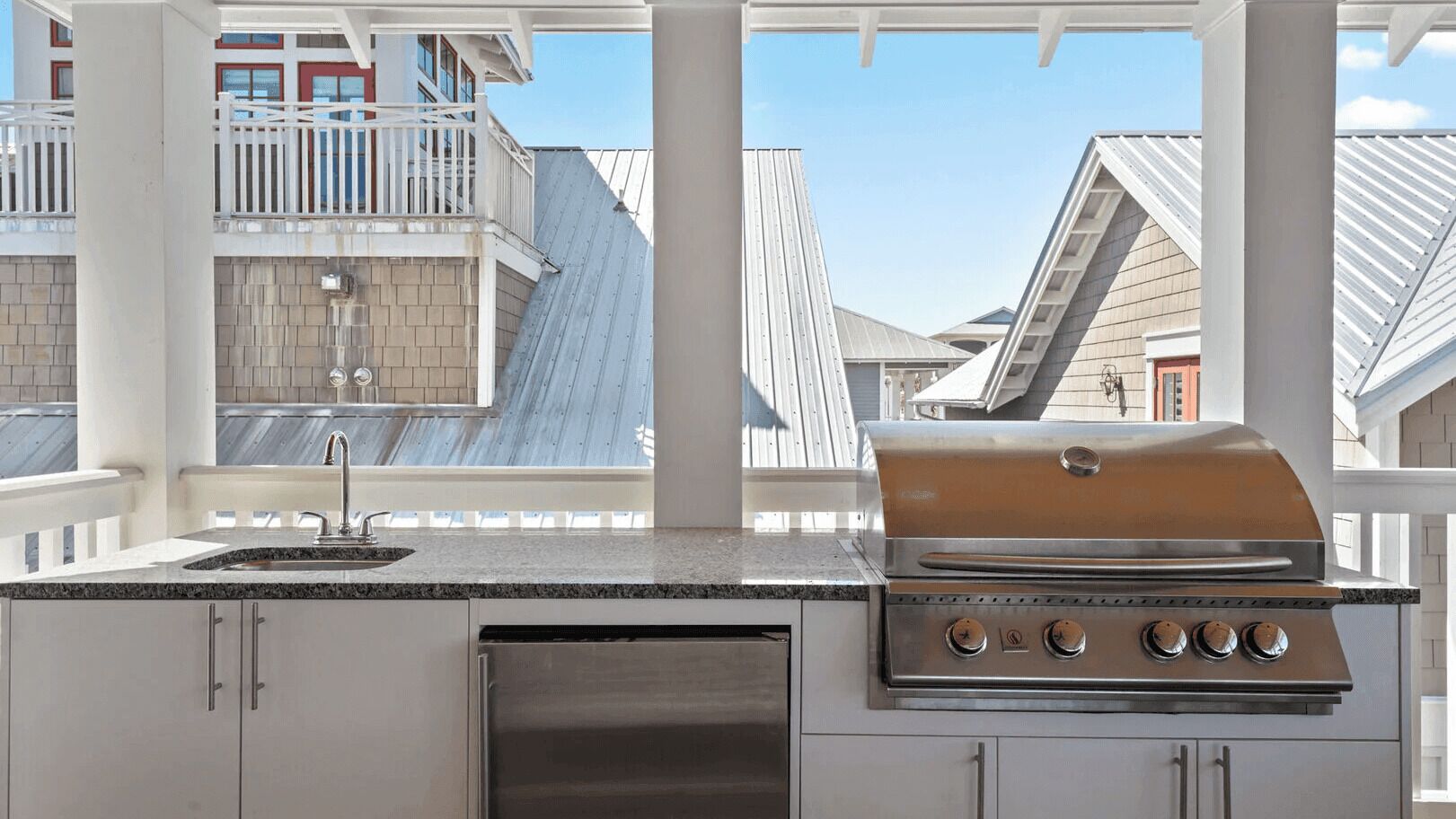 An outdoor kitchen with a grill, a sink, and a dishwasher, set against a backdrop of residential buildings.