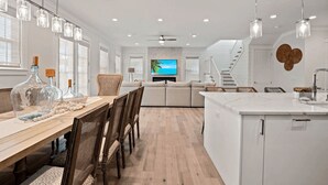 A modern kitchen with a central island, wooden dining table, and chairs. There is a TV mounted on the wall and a staircase leading to another level. - Steps to Beach Pool, Bikes & Game Room Seacrest 5BR (Panama City Beach)