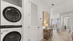 Une buanderie équipée de deux machines à laver et de deux sèche-linge superposés, une console en bois avec une lampe, et un couloir menant à un balcon.
