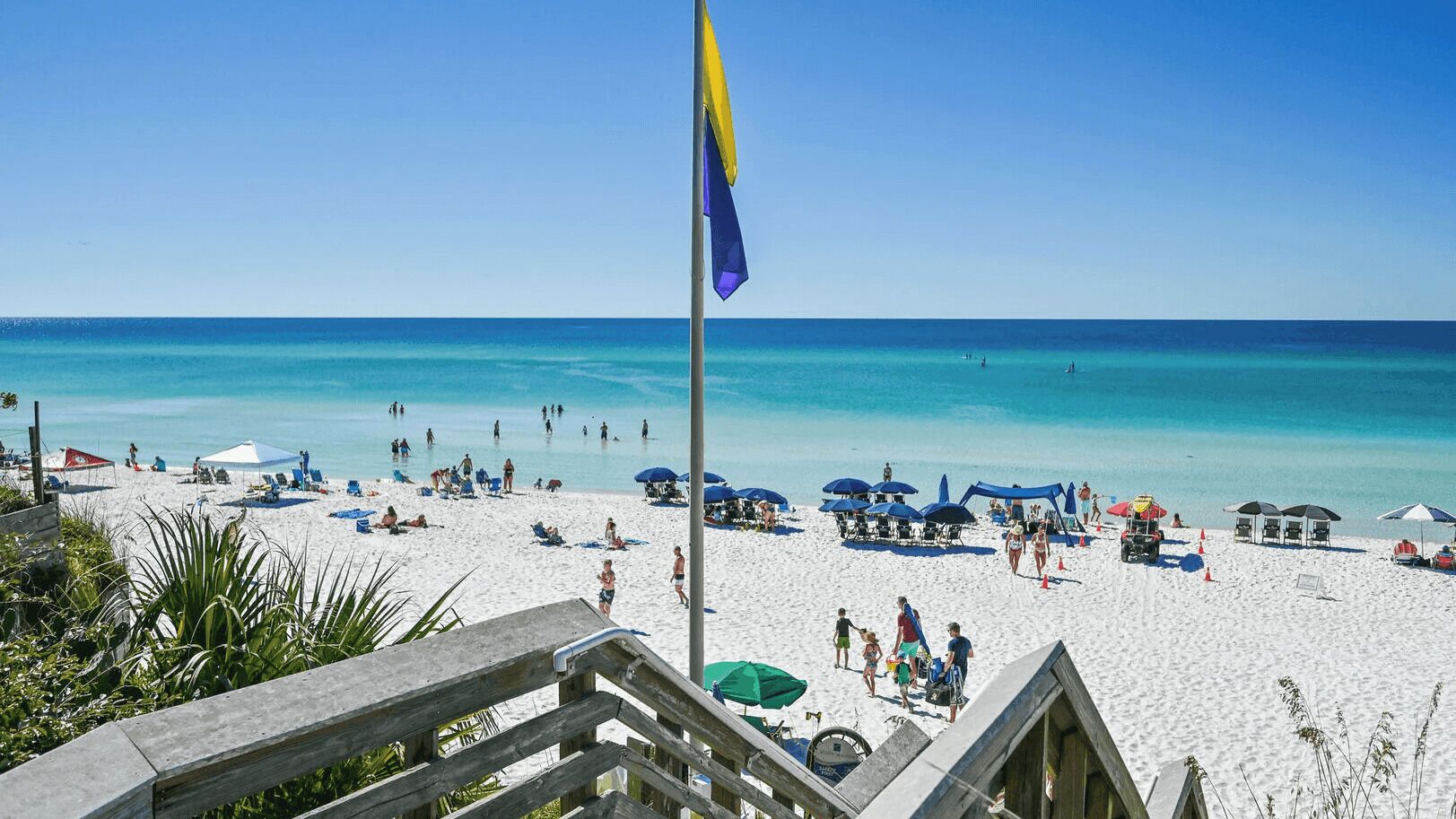 A beach with a flagpole, umbrellas, and people.