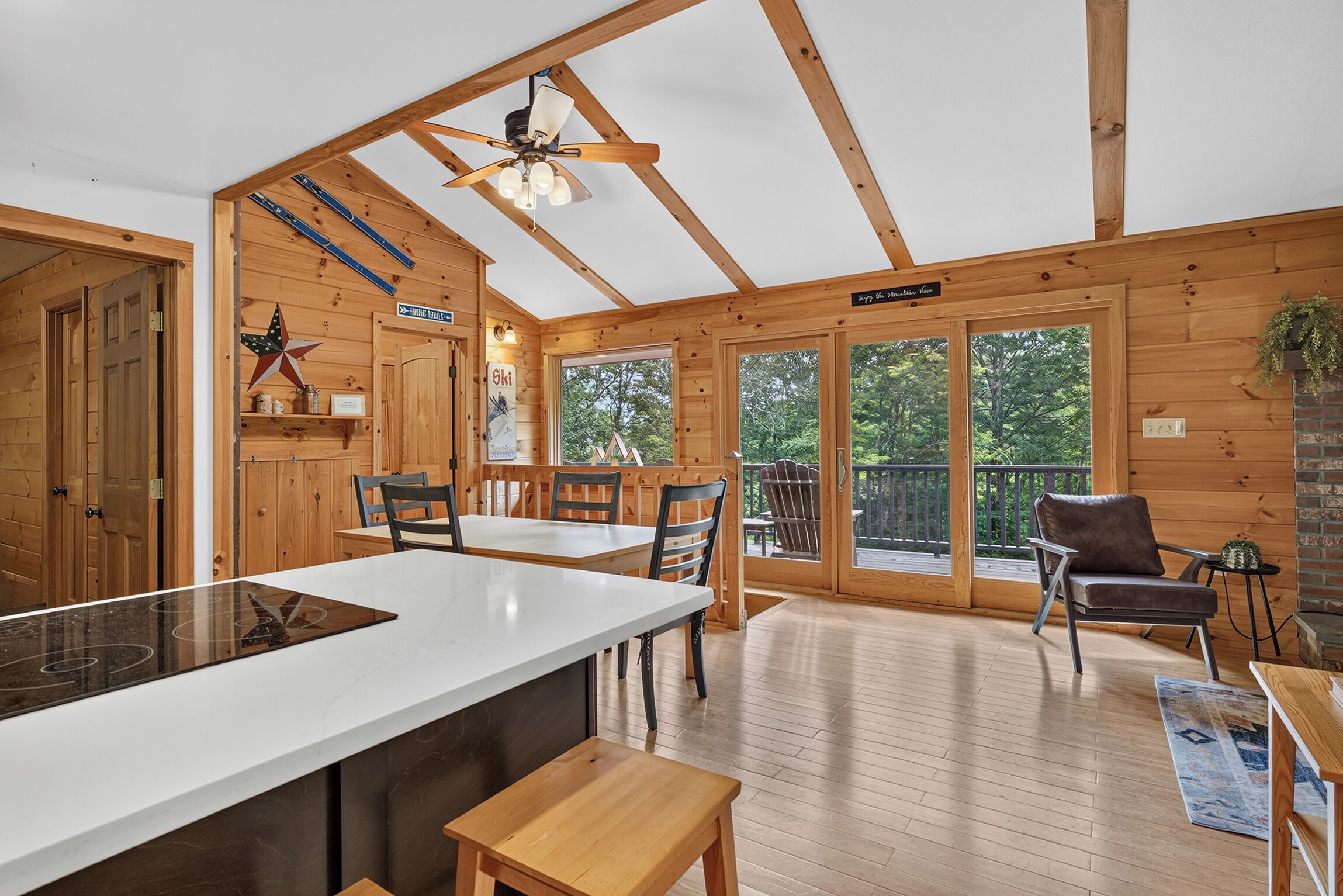 A wooden cabin interior with a kitchen area, dining space, and a balcony with a view of the forest.