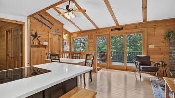 A wooden cabin interior with a kitchen area, dining space, and a balcony with a view of the forest.