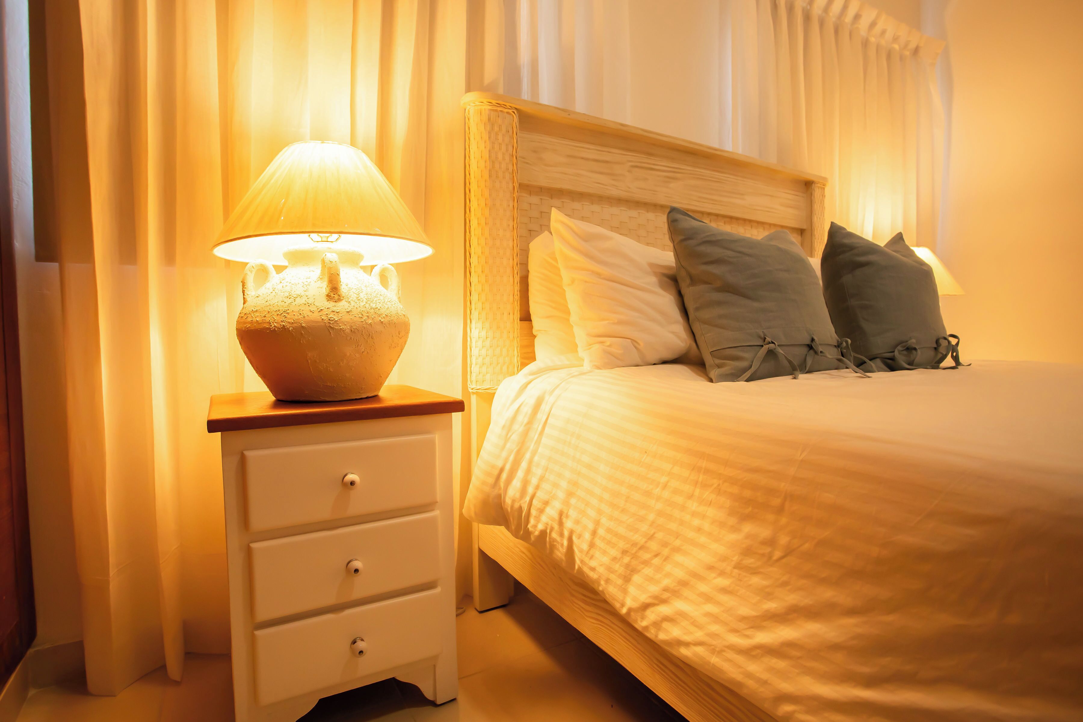 Ein ordentlich bezogenes Bett mit hölzernem Kopfteil, einer Nachttischlampe und einem weißen Nachttisch mit Schubladen.