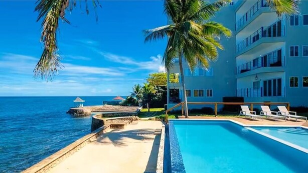 Area piscina di un hotel con vista sull'oceano, palme e un edificio a più piani con balconi.