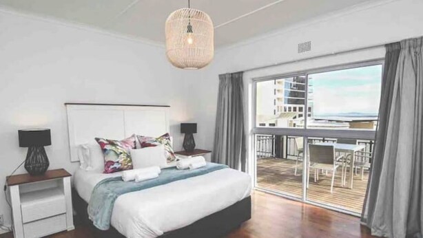 Un dormitorio moderno con una cama grande, una mesita de noche, una lámpara colgante y un balcón con mobiliario exterior.