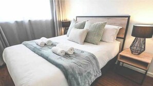 Una cama bien hecha con una manta gris y toallas enrolladas, una mesita de noche de madera con una lámpara, y vistas al exterior a través de una ventana con cortinas.