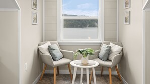 Une pièce d’angle avec deux fauteuils gris, une petite table blanche ornée d’une plante et une fenêtre donnant sur un lac.