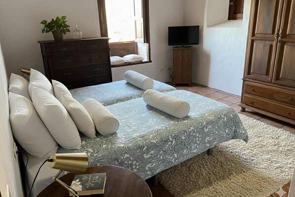 A bedroom with a bed, pillows, a bedside lamp, a dresser, a television, and a window.