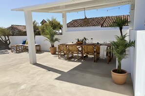 Un patio al aire libre con una mesa, sillas, plantas en macetas y una pared blanca.