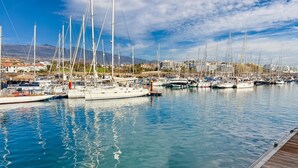 Un puerto deportivo con varios veleros amarrados, un muelle de madera y un telón de fondo de edificios y montañas.