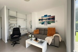 A modern living room with a sofa, coffee table, and a black chair. There is a bookshelf and a window with a view.