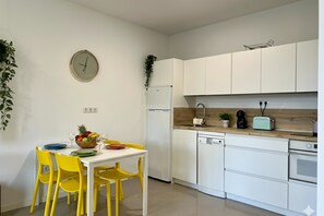 Eine moderne Küche mit weißen Schränken, einem weißen Kühlschrank, einer Holzausziehtafel und einem Essbereich mit Tisch und gelben Stühlen.