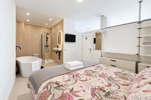 A modern bedroom with a large bed, a built-in bathtub, a vanity with a mirror, and a television.