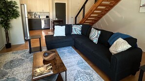 A modern living room with a sectional sofa, a coffee table, and a patterned rug. A staircase with a wooden railing leads to an upper level.