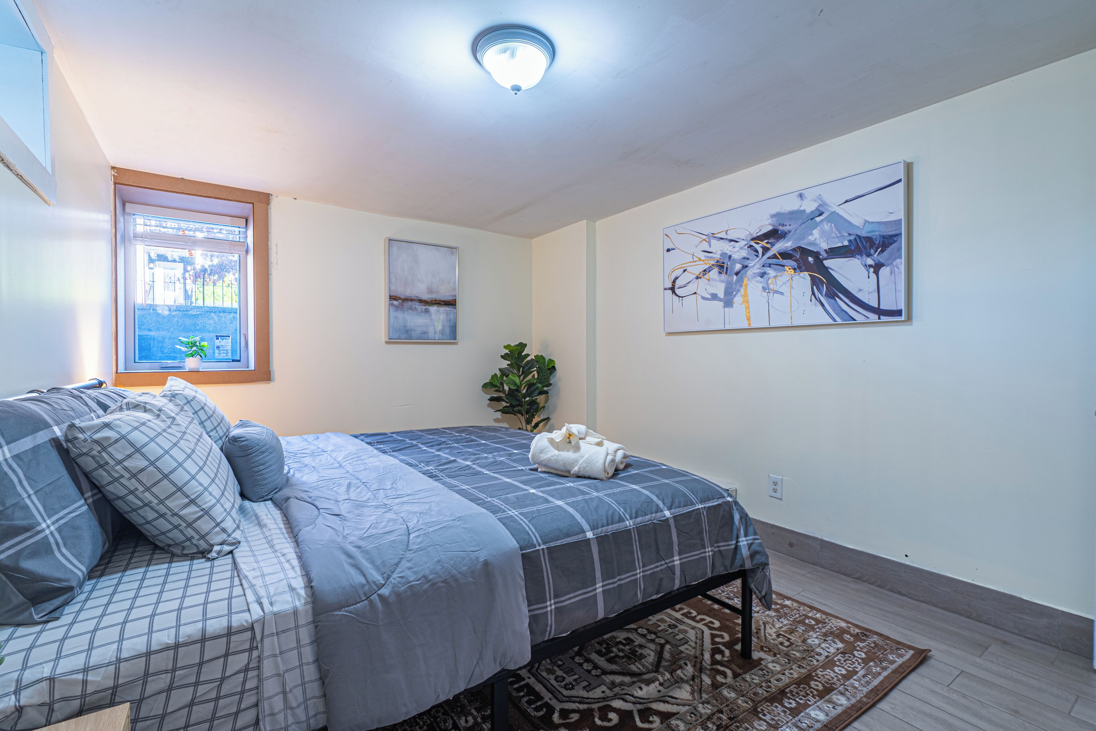 A bedroom with a bed, a window, a plant, and two paintings on the wall.
