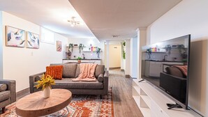 A modern living room with a sofa, coffee table, and TV. There is a kitchen in the background with white cabinets and a countertop. - Adams Village 6-Bed Apt: City Perks, Suburban Vibe (Boston)