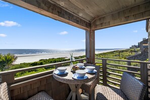 Un balcon en bois avec une table dressée pour le dîner, donnant sur une plage et l’océan.
