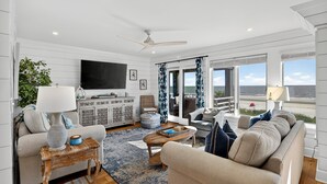 A spacious living room with a large TV, comfortable seating, a wooden coffee table, and a view of the ocean.