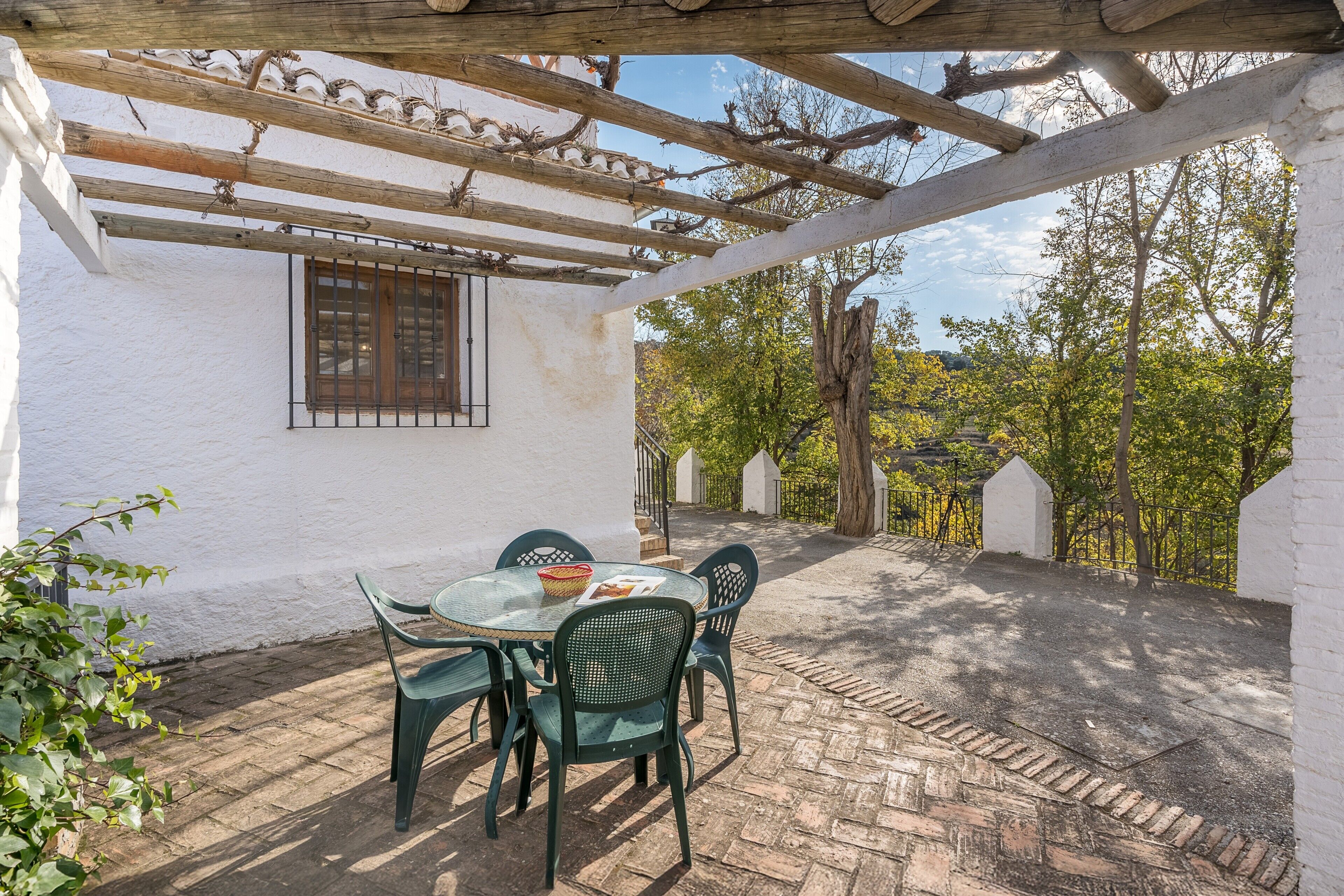 Un patio couvert avec une table et des chaises, une pergola et une vue sur des arbres et un bâtiment blanc.