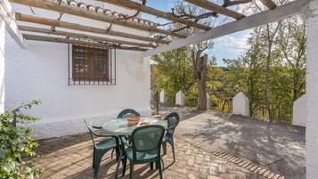 Un patio couvert avec une table et des chaises, une pergola et une vue sur des arbres et un bâtiment blanc.