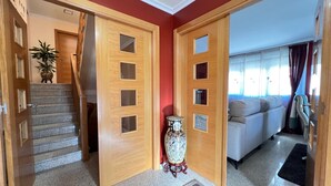 Un pasillo con una escalera, una puerta de madera con paneles de vidrio, un jarrón decorativo y un área de estar con sofá y cortinas.