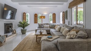A living room with a large sectional sofa, a flat-screen TV mounted on the wall, a fireplace, and a large window with a view of greenery. - Villa Artista (Javea / Xàbia)