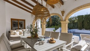 An outdoor patio with a dining table, chairs, and a sofa. There are hanging baskets and a window with a view of greenery. - Villa Artista (Javea / Xàbia)