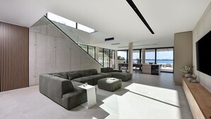 A spacious living area with a large sectional sofa, a coffee table, and a TV on a wooden console. A staircase leads to an outdoor balcony with a view of the sea.