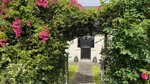 A pathway leading to a building with a black door, surrounded by lush greenery and pink flowers. - Littlemount Farm Cottage (Enniskillen)