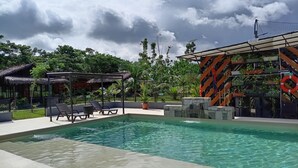 Una piscina con sillas de descanso y un área de estar cubierta, rodeada de vegetación y un edificio con paneles solares.