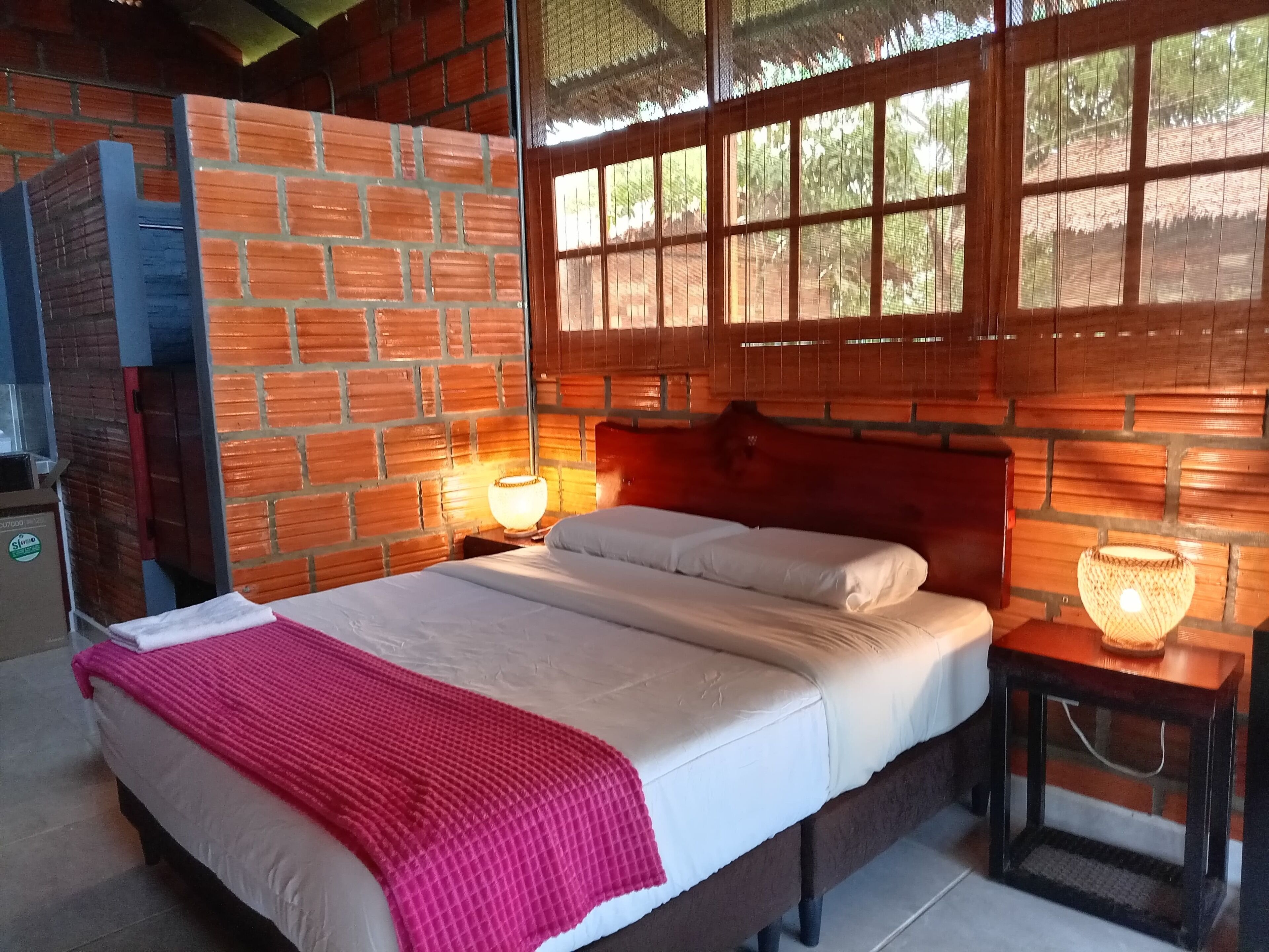 A room with a brick wall, a bed with white bedding and a red blanket, a wooden headboard, and a window with a view of trees.
