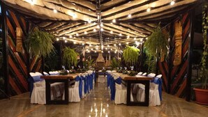 Una larga mesa con sillas blancas y cintas azules, dispuesta en una habitación con techo alto y vigas de madera.