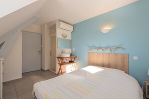 A bedroom with a bed, a wooden headboard, a bedside table with a lamp, a wall-mounted shelf, and a window providing natural light.
