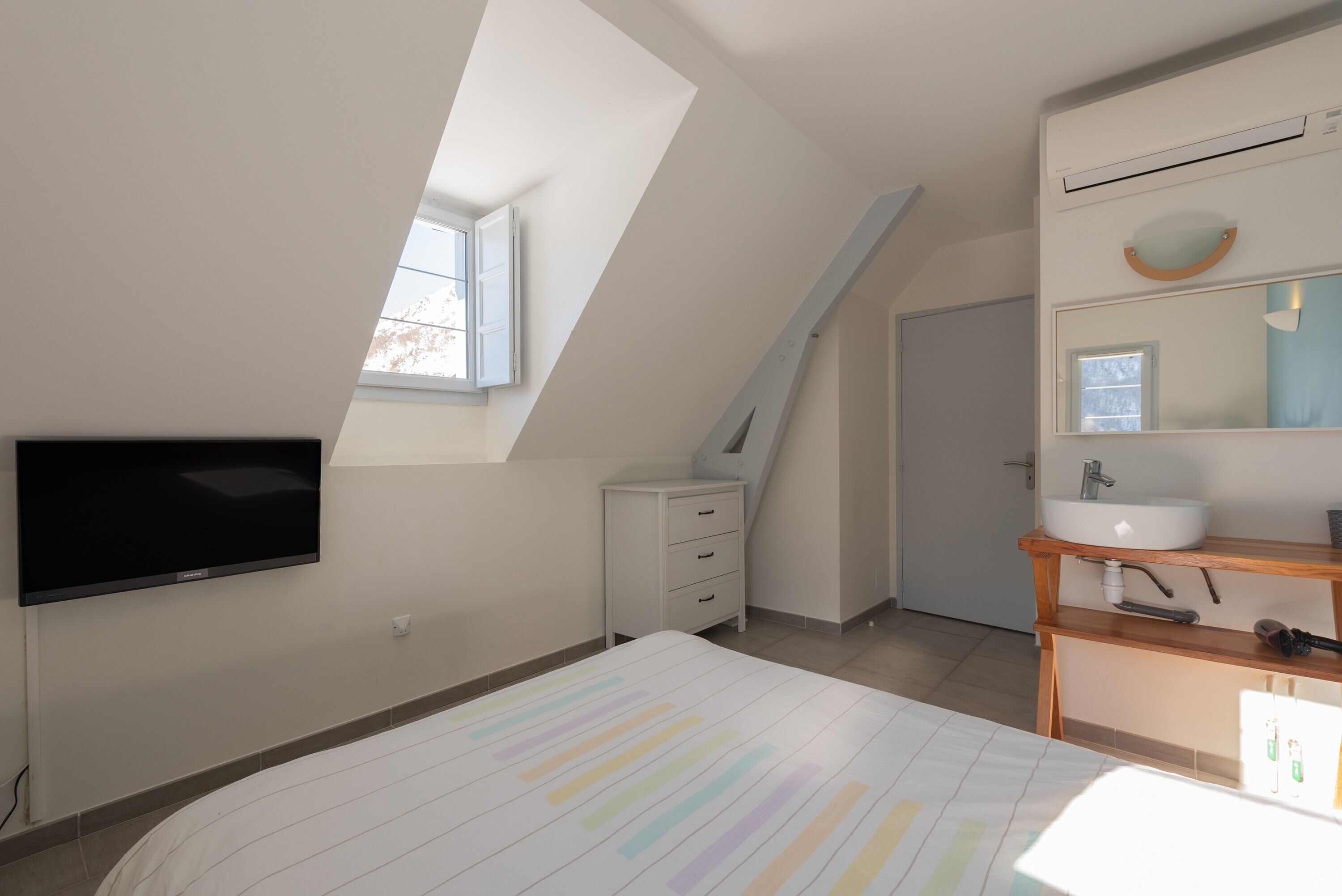 Une chambre avec un plafond en pente, une télévision, une commode, une table de chevet et une fenêtre.