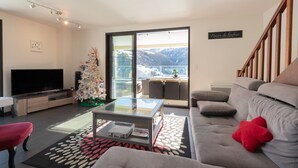 Un saló modern amb televisió de pantalla plana, una taula de centre de vidre, un sofà gris i un coixí vermell. Hi ha un arbre de Nadal i una vista de muntanyes a través d'una gran porta corredissa de vidre.