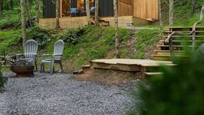 Eine Holzhütte im Wald mit einer Terrasse, zwei Stühlen und einer Feuerstelle.
