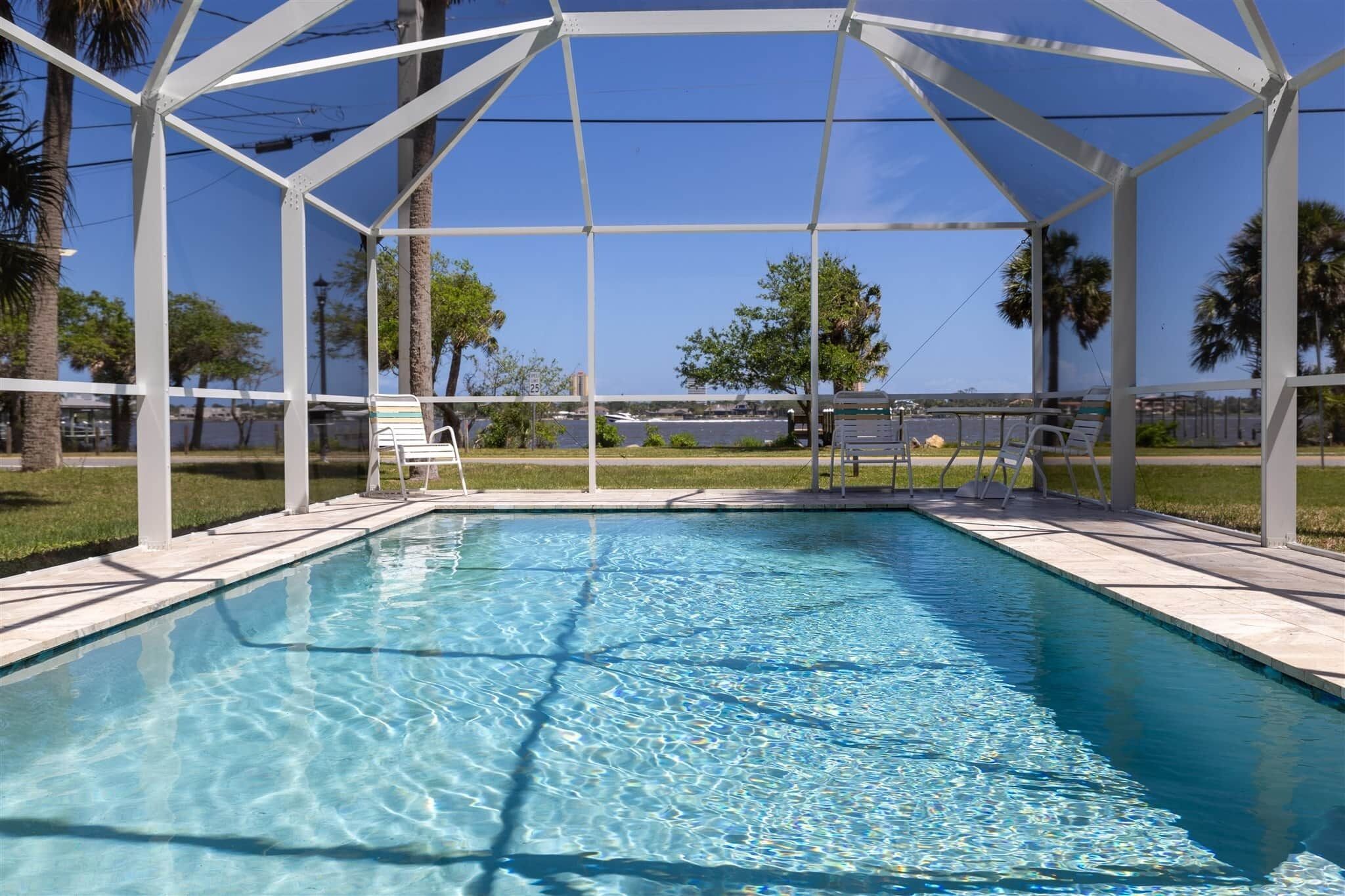 A clear swimming pool with a white roof structure, surrounded by chairs and palm trees.