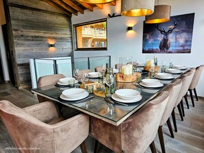 A modern dining room with a long table set for six, featuring glass-top tables, cushioned chairs, and a rustic wooden wall.