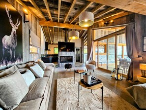 A spacious living room with a wooden ceiling, a fireplace, and a large window overlooking a deck.