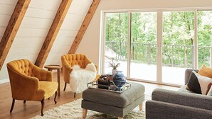 Une pièce mansardée spacieuse avec des poutres apparentes, une grande fenêtre donnant sur l’extérieur et un coin salon confortable agrémenté d’un tapis moelleux et de deux fauteuils.