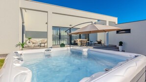 A modern outdoor hot tub area with a glass-enclosed patio, lounge chairs, and a table with a glass of wine. - StunNing home in Nin with sauna (Nin)