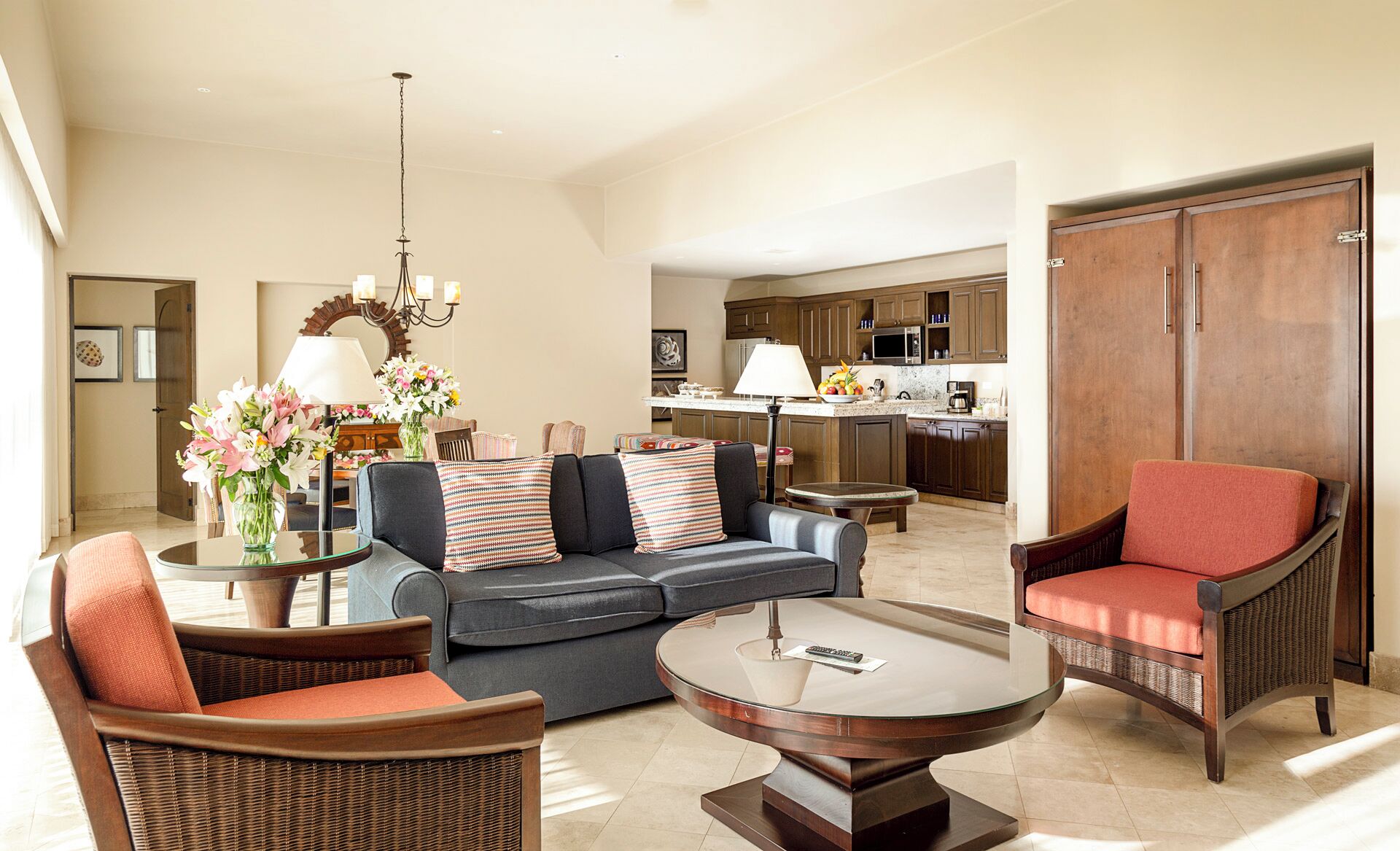 A spacious living room with a sofa, two armchairs, a coffee table, and a dining table. There is a kitchen in the background with wooden cabinets and a microwave.