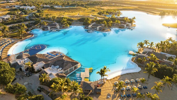 Vue aérienne d’un complexe hôtelier doté d’une grande piscine centrale, de plusieurs villas pour les clients et d’une plage aménagée avec des transats.