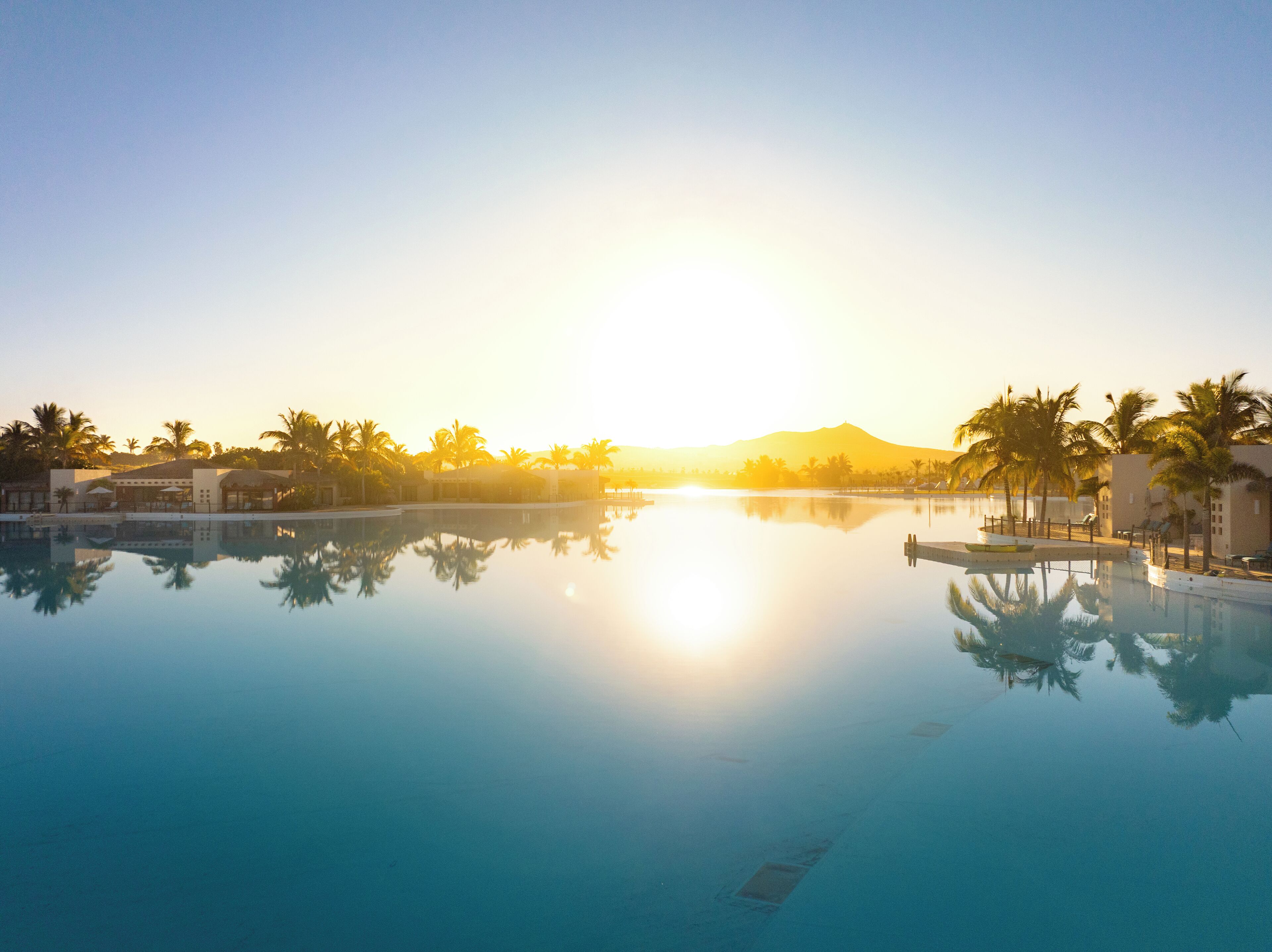 A resort pool with clear water reflecting palm trees and buildings, surrounded by a calm atmosphere.