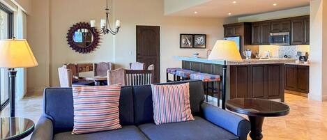 A modern living room with a dark blue sofa, a round table, and a kitchen area with dark wooden cabinets and a granite countertop.