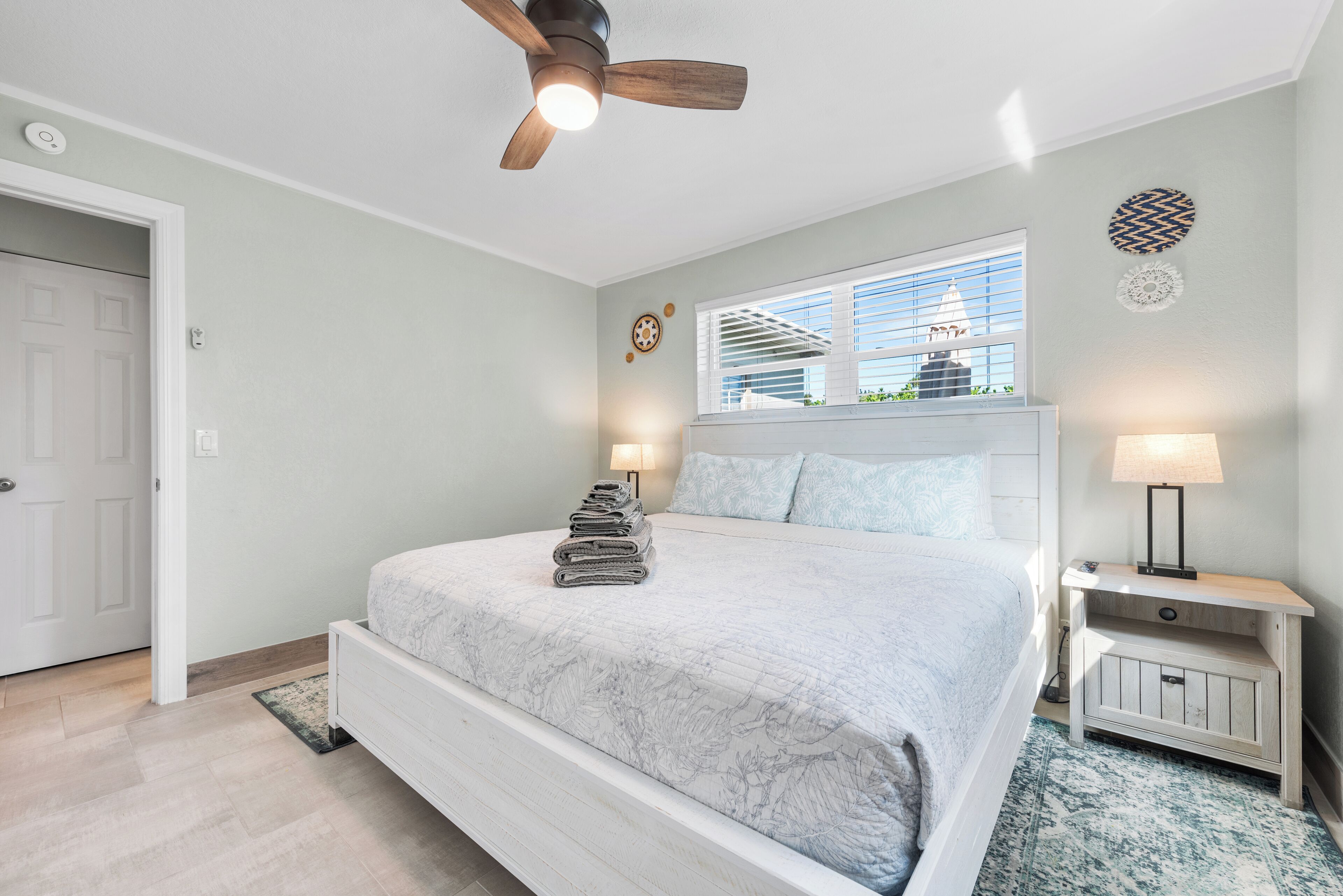 A bedroom with a large bed, a ceiling fan, a window with blinds, a nightstand with a lamp, and a door.