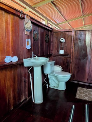 A bathroom with wooden walls, a white sink, two toilets, and a shower area. - Hacienda Río Oro (Puerto Jiménez)